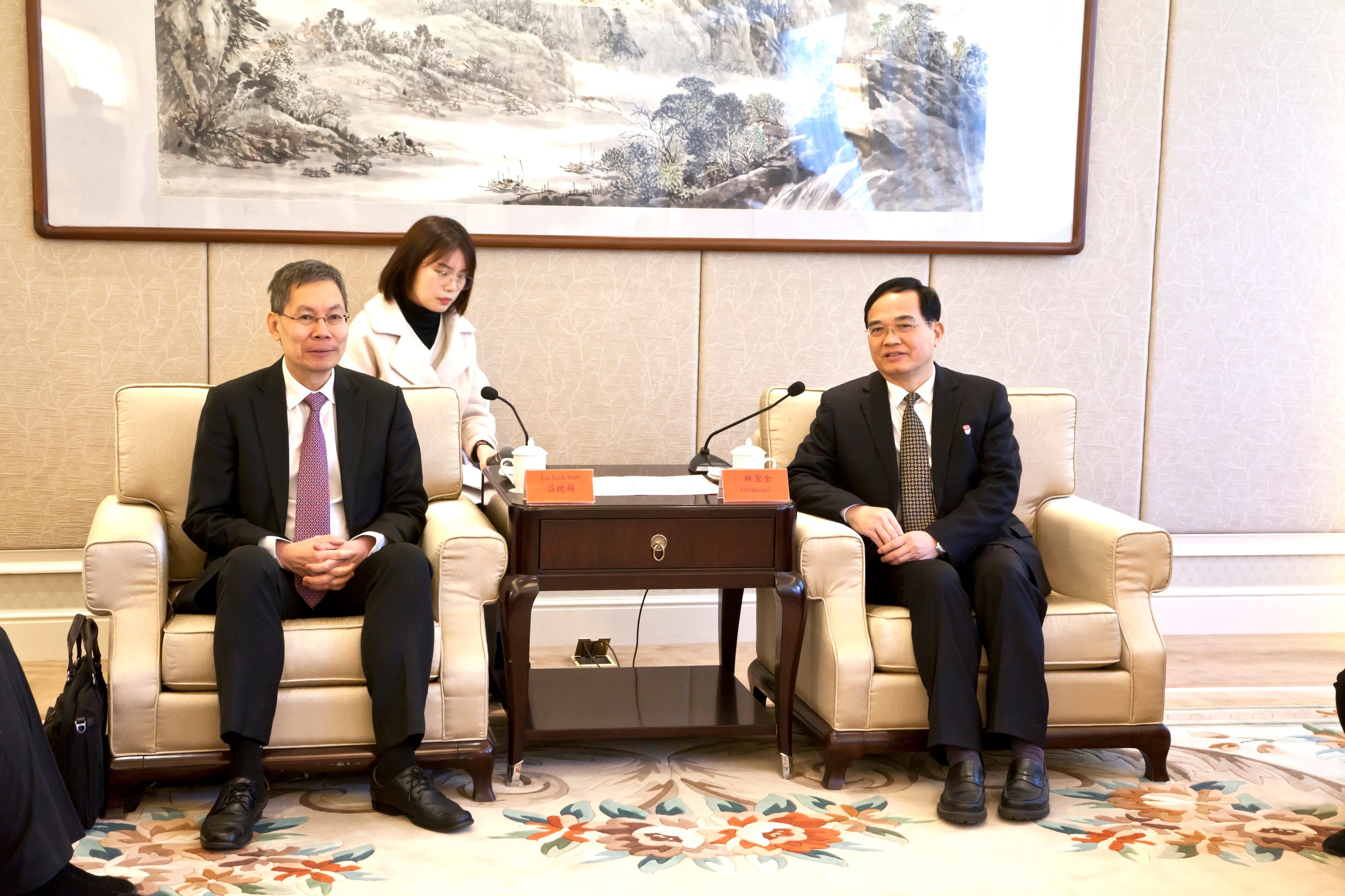 Two men in suits seated, facing forward at a meeting table with microphones. Woman standing behind, touching the table.